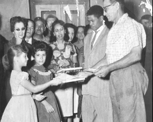 Griselda Aguilera, left at age 7 in 1961, hands literacy certificate to worker she taught to read and write. Aguilera spoke about literacy campaign and Cuban Revolution to hundreds on college campuses, churches and union halls during recent U.S. tour.
