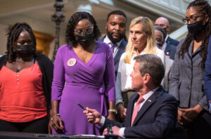 Wanda Cooper-Jones, mother of Ahmaud Arbery, watches as Georgia Gov. Brian Kemp,seated, signed repeal of Civil War-era “citizen’s arrest” law May 10. Three men who killed Arbery claimed law as their defense. Overturn of law was gain of protests against killing.