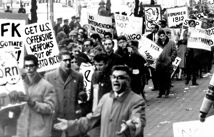 Demonstrations broke out across U.S. and worldwide after Kennedy administration’s announcement of naval blockade against Cuba’s socialist revolution, which took world to brink of nuclear war. Above, protest outside United Nations in New York against threat of Washington invasion of Cuba. Inset, Cuban militia members prepare to defend their revolution arms in hand. Slogan on truck door says, “Standing firm with Fidel.”