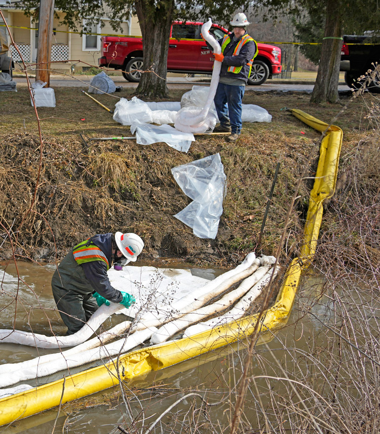 Cleanup crew places booms across stream in East Palestine to try to stem spread of harmful chemicals after Feb. 3 derailment. Norfolk Southern rail bosses, backed by government, ignored impact on residents, setting fire to five cars of vinyl chloride, worsening toxic disaster.