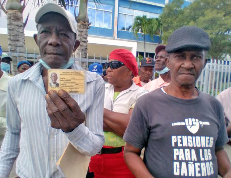 Retired Haitian sugarcane worker Ephesiel Bonel, left, holds up his identification card from formerly state-owned Río Haina Sugar Mill, often only ID these workers have. At right is Yega Fabián, another retired Haitian worker. Shirt says, “Pensions for all sugarcane workers.”