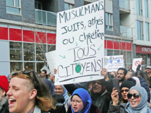 Protest against Law 21 in Montreal, 2019. Placard reads, “Muslims, Jews, atheists and Christians are all citizens.” New Bill 94 expands earlier restrictions on wearing religious symbols at work for all school workers, violating freedoms under federal and Quebec human rights charters.